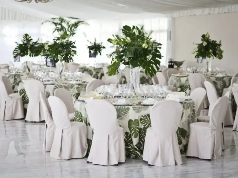 Salón de banquetes con mesas redondas, sillas vestidas de blanco y centros de hojas tropicales